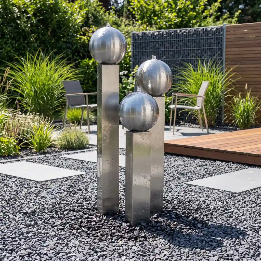 Edelstahl-Gartenbrunnen Balance Sphera 125 cm im 3er-Set, platziert in einem dunklen Kiesbeet neben einer Holzterrasse