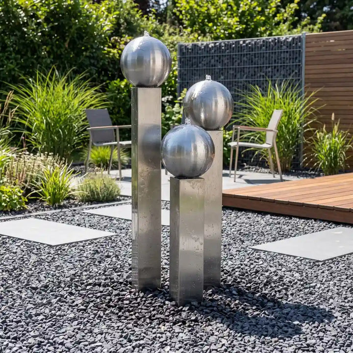 Edelstahl-Gartenbrunnen Balance Sphera 125 cm im 3er-Set, platziert in einem dunklen Kiesbeet neben einer Holzterrasse