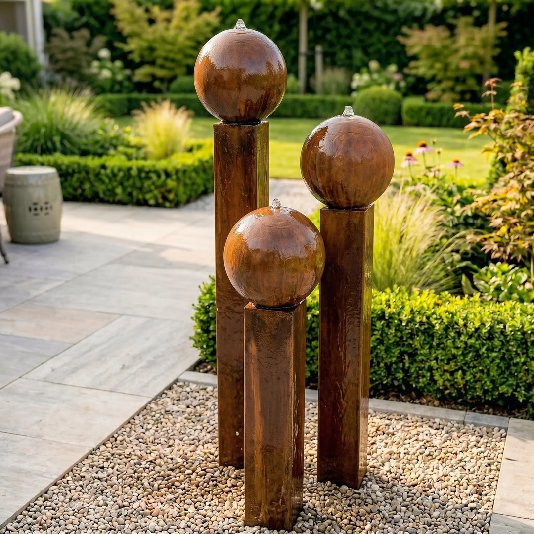 Großer Gartenbrunnen aus Cortenstahl: Das massive 3er Set Komplettset Balance Sphera 125 cm von Gaddia für Präsenz vor grüner Bepflanzung.