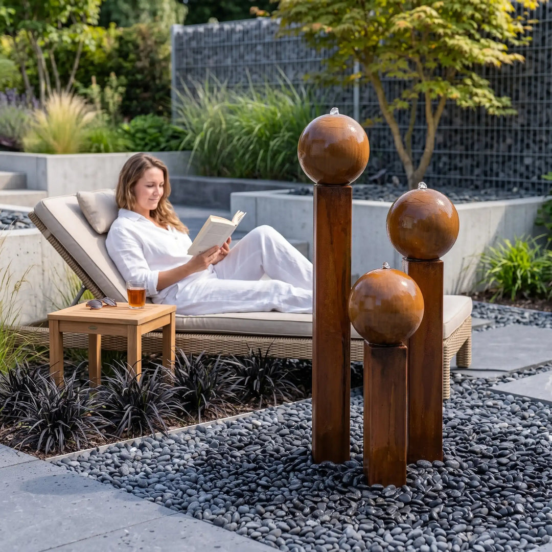 Entspannung am Cortenstahl Gartenbrunnen: Frau liest neben dem sprudelnden 3er Set Komplettset Balance Sphera von Gaddia auf der Terrasse.