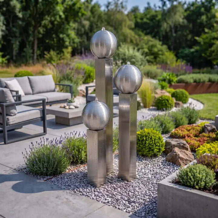 Designbrunnen aus Edelstahl (145 cm) mit drei Kugeln auf grauem Zierkies, platziert vor einer modernen Garten-Lounge